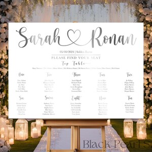 May include: A wedding seating chart on a white sign with the names "Sarah" and "Ronan" in elegant script, with a heart symbol between them. The date "05/10/2024" is displayed. The sign is on a wooden easel, with a floral arch in the background.