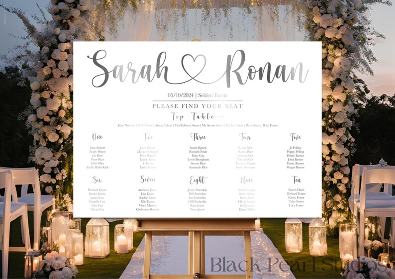 May include: A wedding seating chart on a white sign with the names "Sarah" and "Ronan" in elegant script, with a heart symbol between them. The date "05/10/2024" is displayed. The sign is on a wooden easel, with a floral arch in the background.