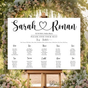 May include: A white wedding seating chart with black calligraphy text. The sign reads "Sarah & Ronan" with a heart symbol, date, and venue. Names are listed under table numbers one through ten. The sign is displayed on a wooden easel.