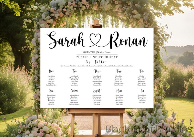 May include: A white wedding seating chart with black calligraphy text. The sign reads "Sarah & Ronan" with a heart symbol, date, and venue. Names are listed under table numbers one through ten. The sign is displayed on a wooden easel.
