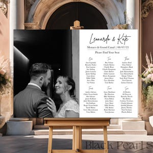 May include: Black and white wedding seating chart with the names Leonardo and Kate at the top. The chart lists guest names in columns labelled "One", "Two", "Three", "Four", and "Five". A black and white photo of the bride and groom is on the left side of the chart.