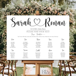 May include: A white wedding seating chart with black calligraphy text. The sign reads "Sarah & Ronan" with a heart symbol, date, and venue. Below, it says "Please Find Your Seat" and lists table numbers and guest names. The sign is displayed on a wooden easel.