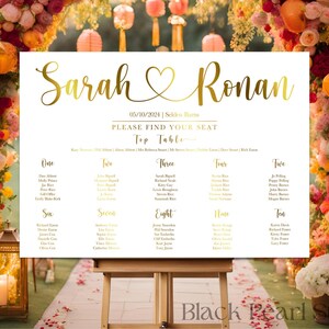 May include: A white wedding seating chart with gold lettering, displaying the names "Sarah & Ronan" and the date. The chart is on an easel, surrounded by floral arrangements and lanterns.