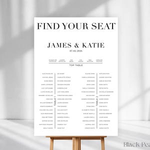 May include: A white wedding seating plan displayed on a wooden easel. The text at the top reads "FIND YOUR SEAT", followed by "JAMES & KATIE" and the date "07.02.2026". Guest names are listed below, organised by table.