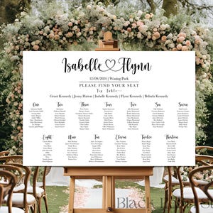 May include: A white wedding seating chart on an easel, with the names "Isabelle Flynn" and the date "12/09/2024" at the top. The chart lists guest names under table numbers, with a floral arch in the background.