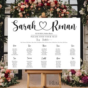 May include: A wedding seating chart with the names "Sarah" and "Ronan" in elegant script, separated by a heart symbol. The date "05/10/2024" is displayed, along with the venue. The chart is on a wooden easel, surrounded by floral arrangements.