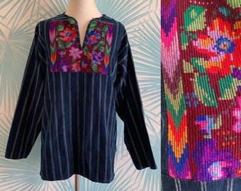 Puede incluir: Una camisa de manga larga a rayas azules y blancas con detalles florales bordados de colores en el cuello. El bordado presenta una variedad de colores, incluyendo rojo, rosa, morado, verde y amarillo.