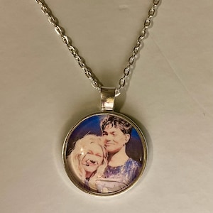 May include: Silver necklace with a circular pendant displaying a colour portrait of two individuals. The pendant has a silver edge and a chain composed of small, connected links.
