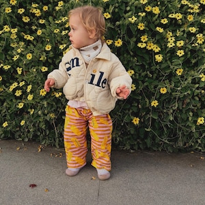 May include: A child wearing a cream-colored quilted jacket with the word "smile" and a matching scarf. The child is also wearing striped pants with a banana pattern and pink shoes, standing in front of a hedge with yellow flowers.