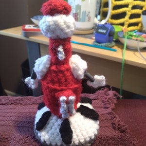 May include: A crocheted robot toy with a red body, white arms and legs, and a black and white base.