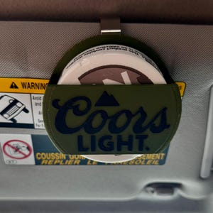 May include: A green car visor organizer with the Coors Light logo in navy blue. The organizer holds a white container with a brown lid. The visor is gray and has a warning label.