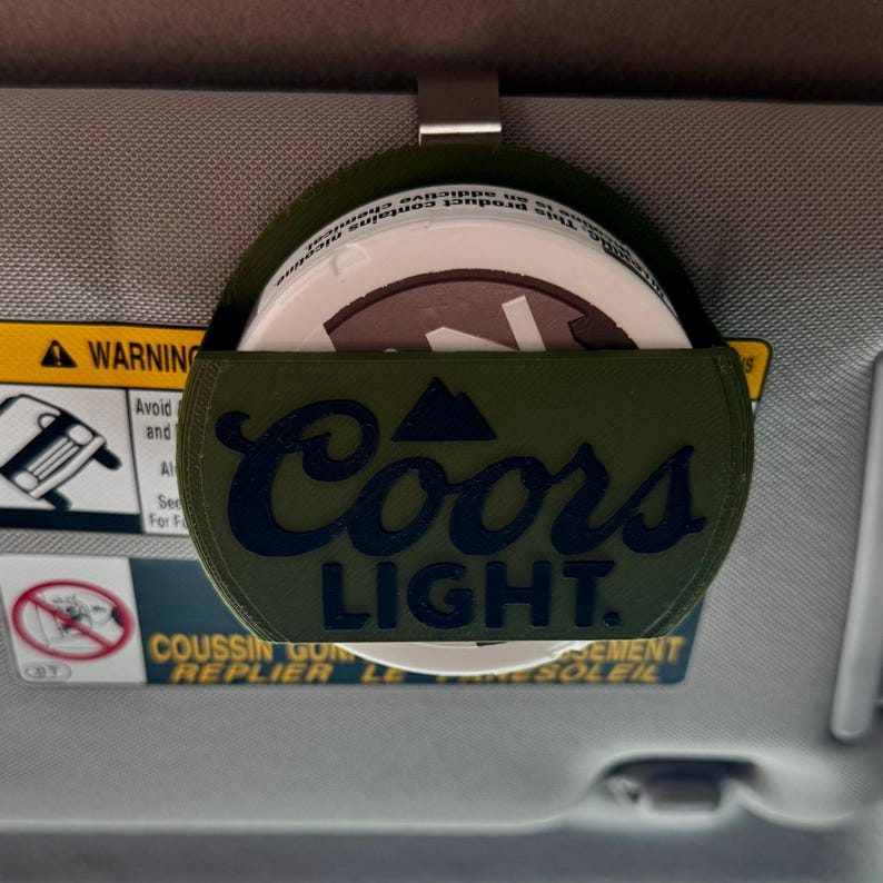 May include: A green car visor organizer with the Coors Light logo in navy blue. The organizer holds a white container with a brown lid. The visor is gray and has a warning label.