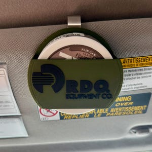 May include: An olive green visor organizer with a metal clip, holding a round container. The organizer features the text "RDO EQUIPMENT CO." in black. The container has the text "FLIP CAN CLAIM REWARD".