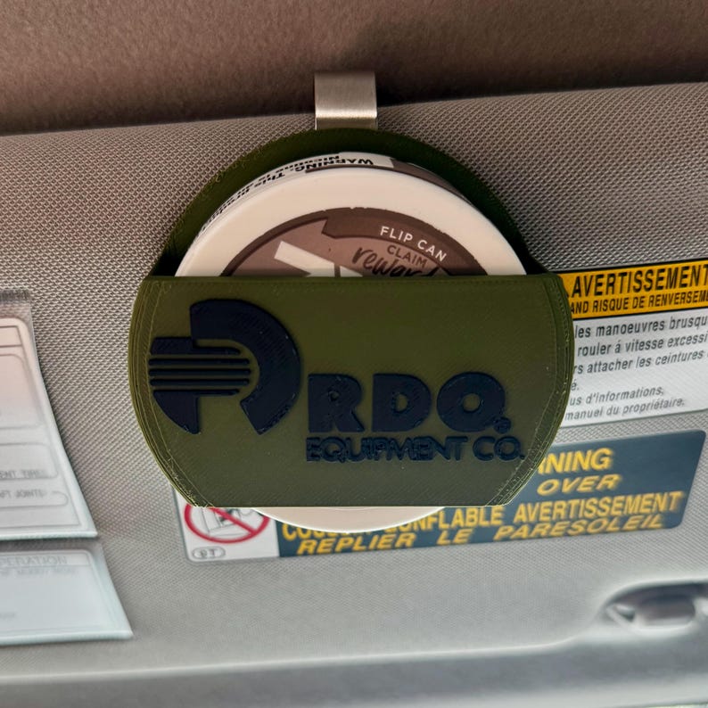 May include: An olive green visor organizer with a metal clip, holding a round container. The organizer features the text "RDO EQUIPMENT CO." in black. The container has the text "FLIP CAN CLAIM REWARD".