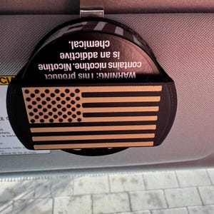 May include: A black car visor organizer with a metal clip. The organizer features a laser-cut American flag design in tan and black. The text "WARNING: This product contains nicotine. Nicotine is an addictive chemical" is printed on the top.