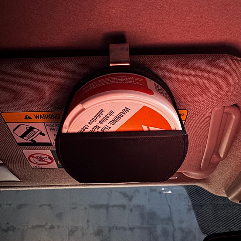 Peut inclure: Une pochette en cuir noir avec un clip en m&eacute;tal, con&ccedil;ue pour contenir un r&eacute;cipient rond. Le r&eacute;cipient est blanc et orange, avec l'avertissement "WARNING: This product contains nicotine. Nicotine is an addictive chemical."