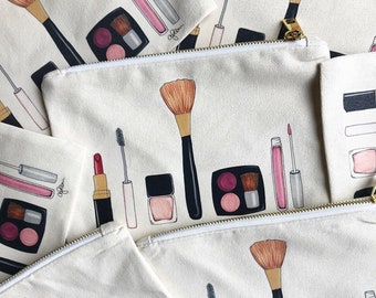 May include: A set of five white canvas makeup bags with gold zips. Each bag features a different design of makeup brushes, lipstick, eyeshadow palettes, and mascara. The bags have the word "Glow" printed on them.