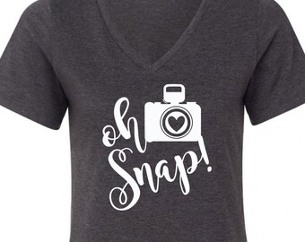 May include: A dark gray V-neck t-shirt with a white graphic of a camera with a heart in the viewfinder and the text "oh snap!" in a cursive font.