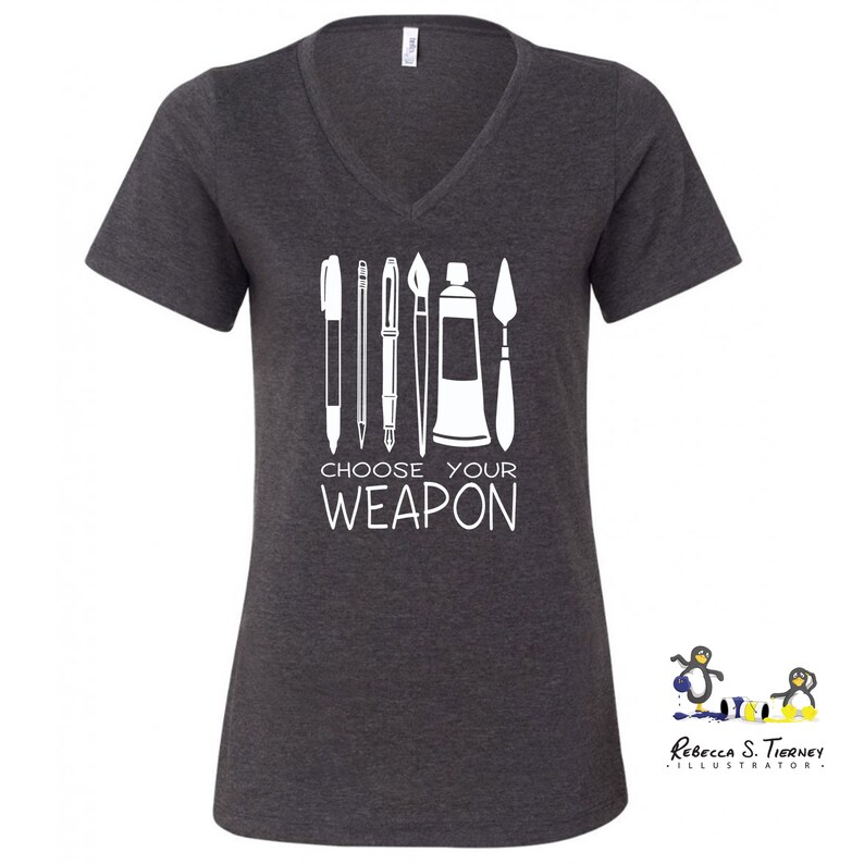 May include: A dark heather grey V-neck t-shirt with a white graphic of art supplies, including paintbrushes, pens, a paint tube, and a palette knife. The text "Choose Your Weapon" is printed below the graphic.