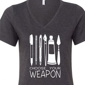 May include: A dark gray V-neck t-shirt with a white graphic of art supplies, including a pen, pencil, paintbrush, paint tube, and a palette knife. The text "Choose Your Weapon" is printed below the graphic.
