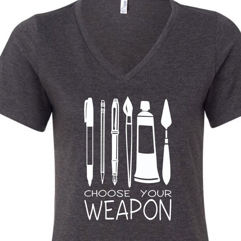 May include: A dark gray V-neck t-shirt with a white graphic of art supplies, including a pen, pencil, paintbrush, paint tube, and a palette knife. The text "Choose Your Weapon" is printed below the graphic.