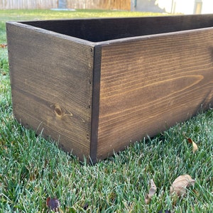 May include: A dark brown wooden planter box with a natural wood grain. The planter is sitting on a bed of green grass.