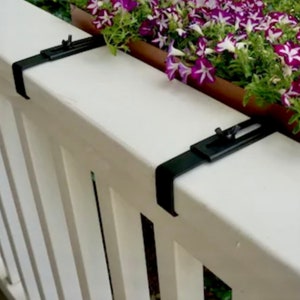 May include: Two black metal plant brackets mounted on a white wooden fence. The brackets are designed to hold a planter box.