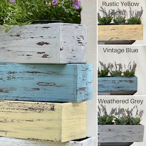 May include: Three stacked wooden planter boxes in various distressed paint colors: white, blue, and yellow. The boxes are labeled "Rustic Yellow", "Vintage Blue", and "Weathered Grey".
