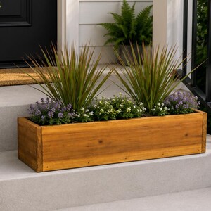 May include: A rectangular wooden planter box filled with green and purple flowering plants. The box is made of light brown wood and sits on a concrete step. The plants have long, thin leaves and small, colorful flowers.
