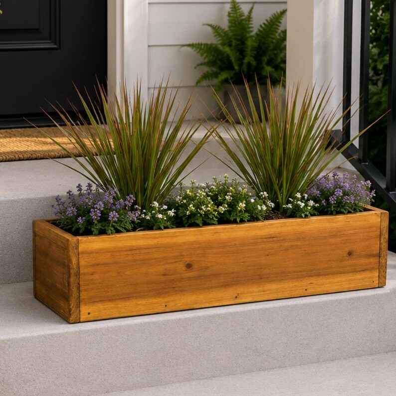 May include: A rectangular wooden planter box filled with green and purple flowering plants. The box is made of light brown wood and sits on a concrete step. The plants have long, thin leaves and small, colorful flowers.