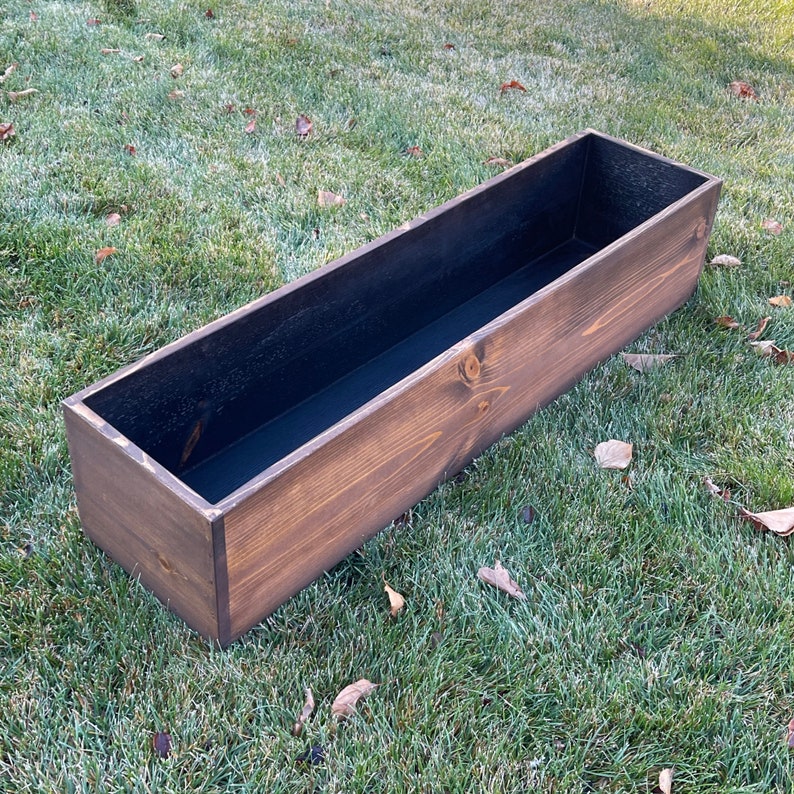 May include: A long, rectangular wooden planter box with a dark brown stain. The planter has a black liner inside.