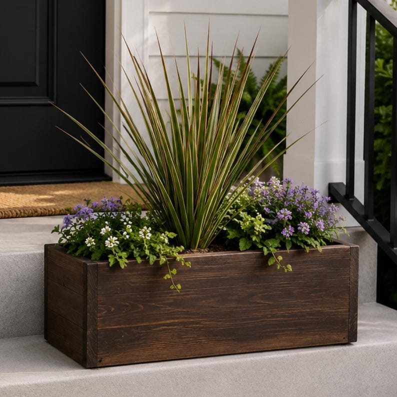 May include: A rectangular, dark stained wooden planter box filled with various plants and flowers. The box sits on a concrete step, with a black door and railing in the background. The plants include tall, spiky green and brown leaves, and small purple and white flowers.