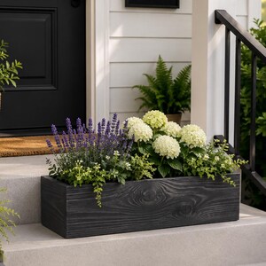 May include: A rectangular, dark gray wooden planter box filled with lavender, white hydrangeas, and greenery. The planter sits on a concrete step in front of a white-sided house with a black door.