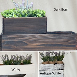 May include: Three wooden planter boxes in different finishes: dark burn, white, and antique white. Each planter box is filled with green plants and purple flowers.
