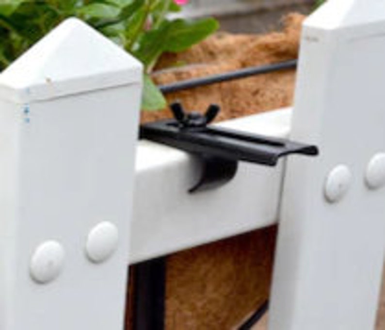 May include: Black metal plant hanger bracket with a white plastic fence post in the background. The bracket has a screw and a nut to secure it to the fence post.