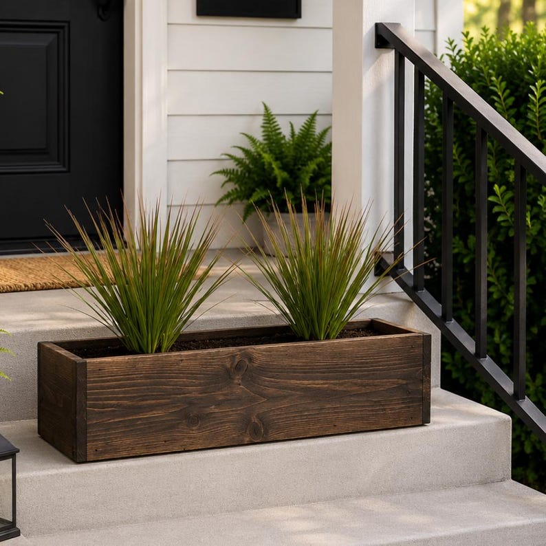 May include: A rectangular, dark stained wooden planter box filled with two ornamental grass plants. The planter sits on a concrete step, with a black door and white siding in the background. The plants have long, thin, green blades.