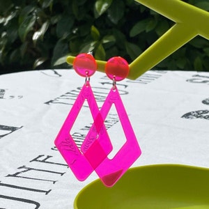 May include: A pair of neon pink diamond-shaped earrings with a hollow centre. The earrings are hanging from a green display stand.