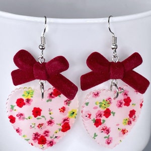 Op de afbeelding: Een paar hartvormige oorbellen met een bloemenprint en een bordeauxrode fluwelen strik aan elke oorbel.