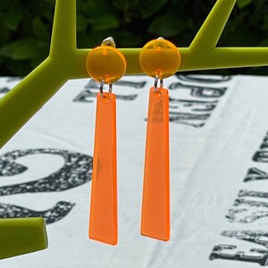 May include: A pair of orange dangle earrings with a round, orange top. The earrings are made of acrylic and have a silver hook.