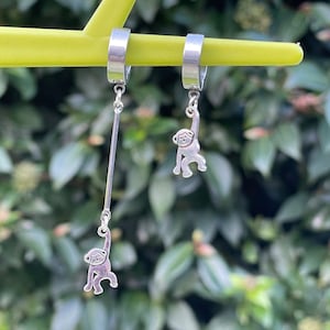 Può includere: Un paio di orecchini d'argento a forma di scimmia. Ogni orecchino presenta un piccolo ciondolo a forma di scimmia appeso a un cerchio d'argento. I ciondoli a forma di scimmia pendono da una catena d'argento.