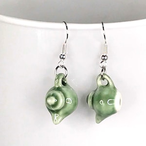 May include: A pair of green ceramic teapot earrings with silver hooks. The teapots have white accents and are a whimsical design.