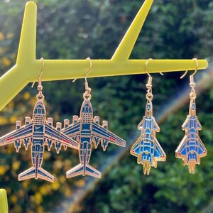 Può includere: Due paia di orecchini pendenti con design a forma di aereo. Il primo paio raffigura grandi aerei di linea blu scuro con contorni dorati. Il secondo paio mostra caccia militari azzurri più piccoli con dettagli in metallo dorato e ganci.