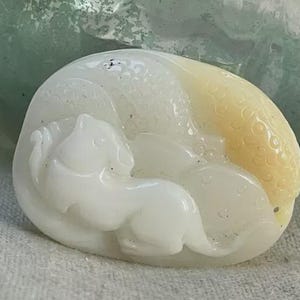 May include: A white, oval-shaped carving of a sleeping cat, with a yellow accent on the right side. The cat is curled up, and the carving has a smooth, polished finish. The background is a blurred green and white.