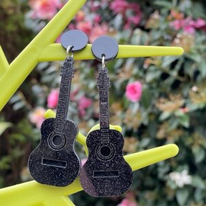 May include: A pair of black glitter guitar earrings with black circular posts. The earrings are hanging from a yellow branch.