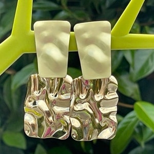 May include: A pair of gold-toned earrings with a textured, irregular shape. The earrings are hanging from a green branch.