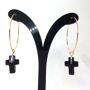 May include: A pair of gold hoop earrings with black cross charms hanging from them. The earrings are displayed on a black earring stand.