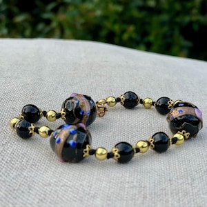 May include: A beaded bracelet featuring black and gold beads. The bracelet has three larger, round, black beads with gold and blue accents. The smaller beads are black and gold, strung together with black cord.