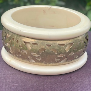 Puede incluir: Un cuenco decorativo redondo de color crema con una banda de filigrana de latón. El cuenco tiene una parte superior ancha y abierta y un diseño calado decorativo alrededor del centro. La base también es de color crema y redonda.