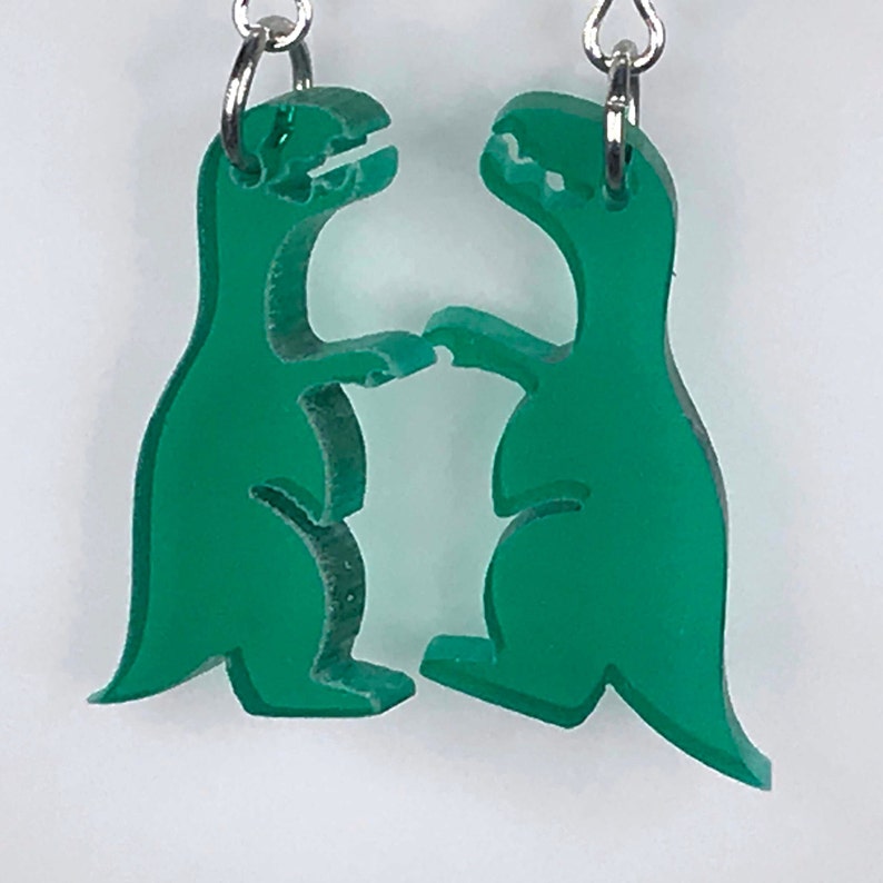 May include: A pair of green dinosaur earrings with silver hooks. The dinosaurs are facing each other and appear to be holding hands.