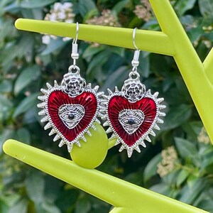 May include: A pair of silver earrings with red heart-shaped charms. Each charm features a silver eye with a black pupil and a silver floral design at the top.